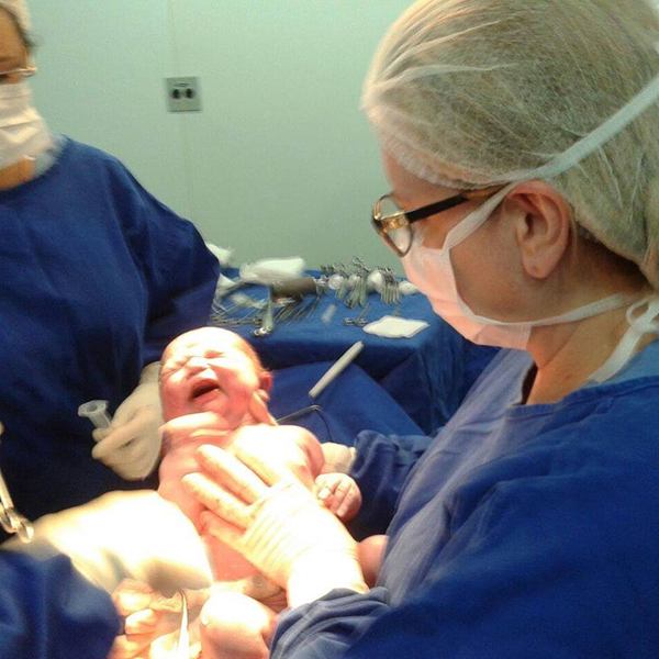 Dra. Lilian em um centro cirúrgico, vestindo roupa zaul cirúrgica, segurando um bebê rescem-nascido e acompanhada por sua assistente.