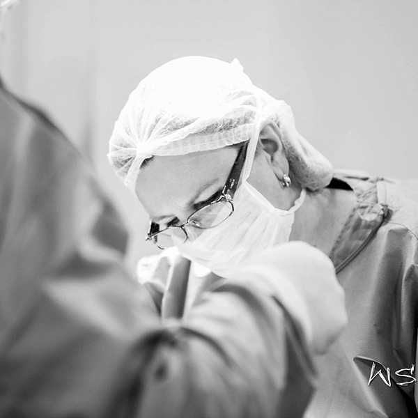 Dra. Lilian no centro cirúrgico de oculos e roupa cirurgica realizando uma cirurgia.