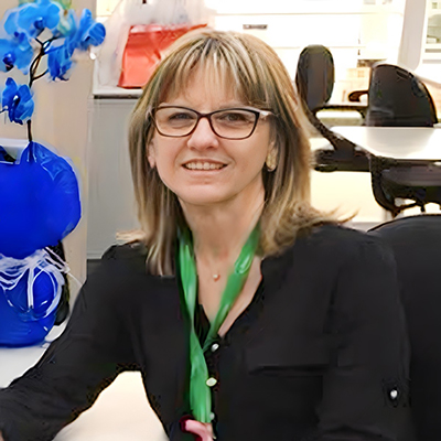 Dra. Lilian sentada, sorrindo e olhando para a câmera, vestindo roupa preta e usando óculos.