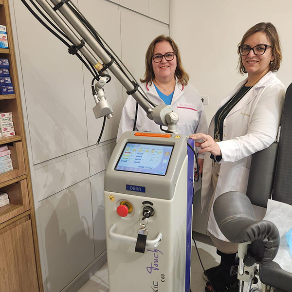 Dra. Joseane acompanhada de sua colega, elas estão em uma sala sorrindo e olhando para a câmera enquanto apresentam o equipamento a laser possivelmente utilizado com suas pacientes.
