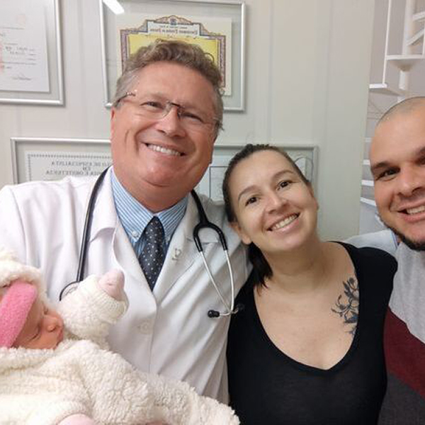 O Dr. Carlos Rogério segura um bebê nos braços, ao lado dos pais jovens, todos sorrindo e olhando para a câmera em um momento de alegria e realização. A cena se passa no consultório do Dr. Carlos Rogério, onde uma parede ao fundo exibe quadros e diplomas em molduras discretas sobre uma superfície branca, reforçando o ambiente acolhedor e profissional.