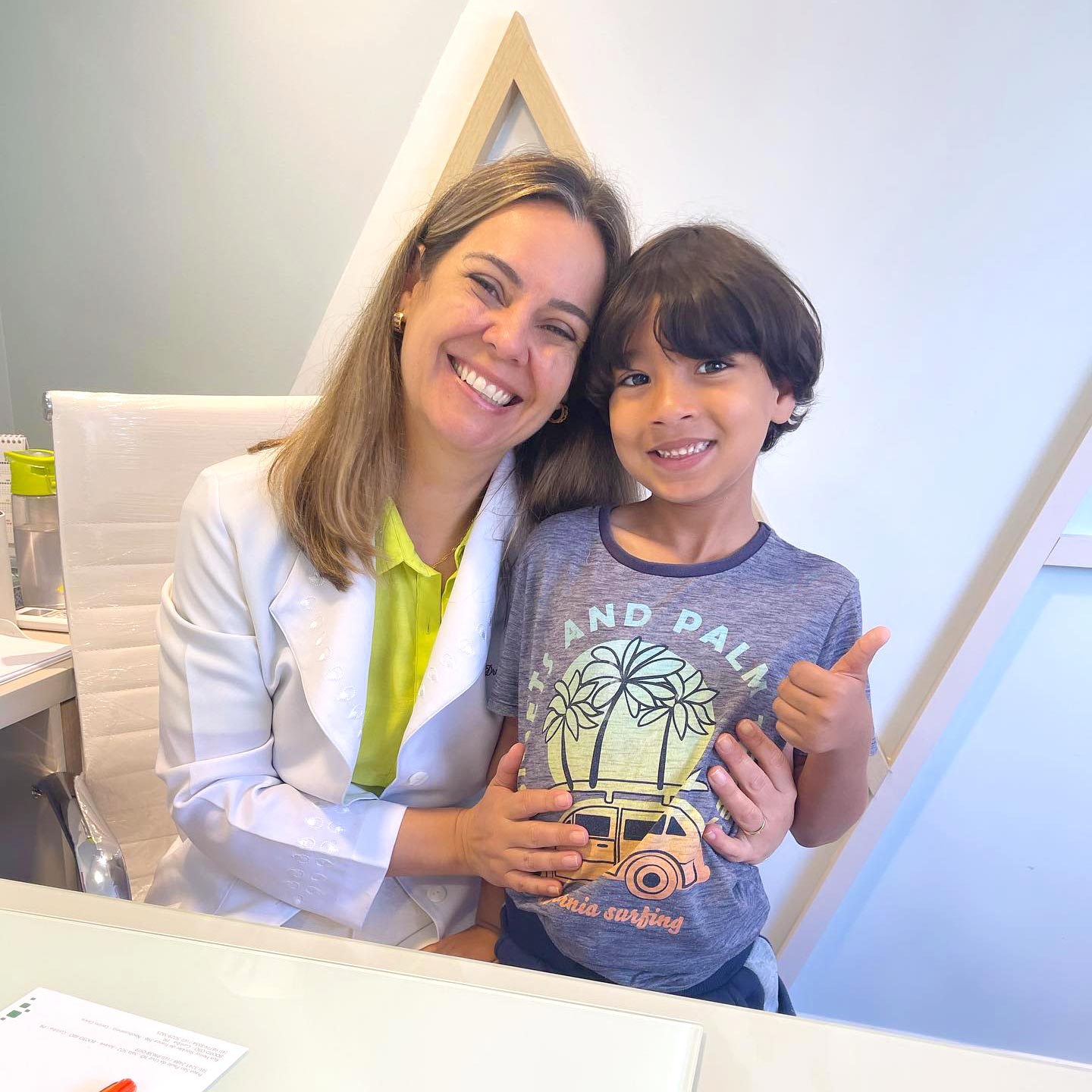 Dra. Luciana em seu consultório sentada em sua mesa e abraçando um pequeno paciente menino ainda criança, ambos sorrindo e olhando para a câmera.