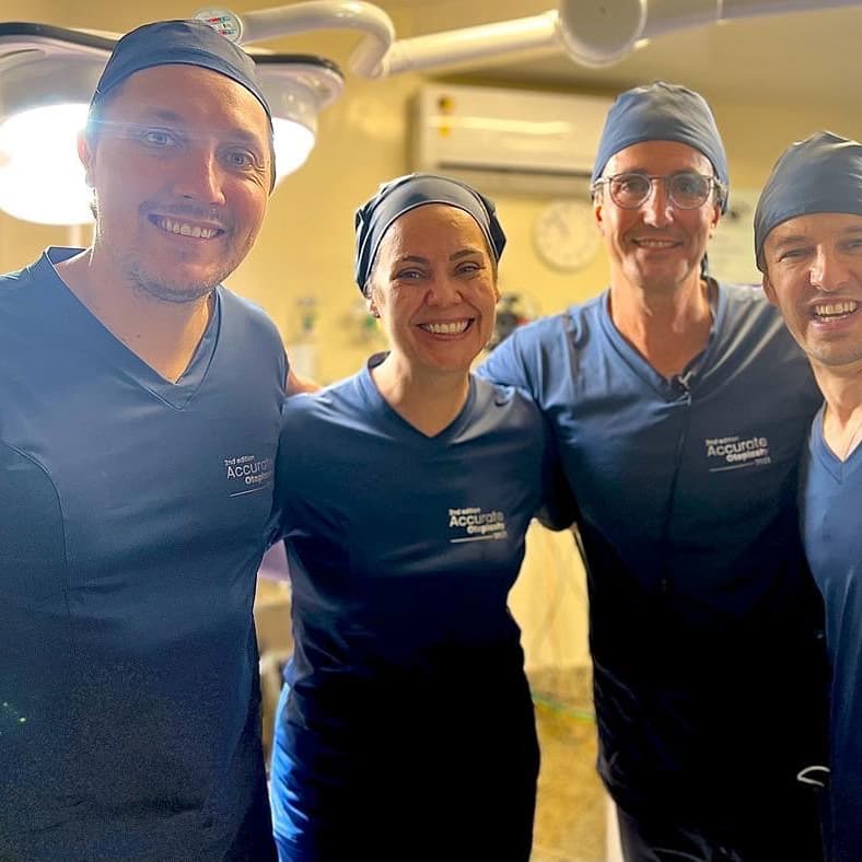 Dra. Luciana em um centro cirúrgico com outros quatro colegas homens, todos abraçados, sorrindo e olhando para câmera, todos em uniforme azul cirúrgico.