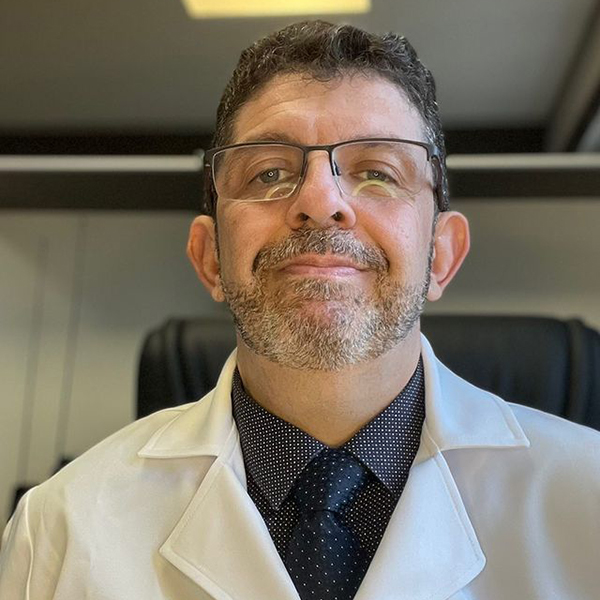O Dr. Marcelo está em seu closet, olhando diretamente para a câmera com a cabeça erguida e um suave sorriso nos lábios, transmitindo confiança e serenidade. A expressão dele é tranquila, refletindo uma presença acolhedora e simpática.