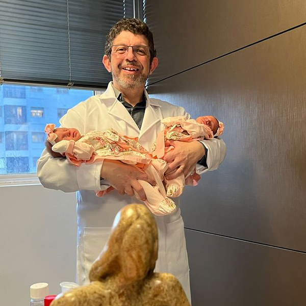 Dr. Marcelo está em seu consultório, vestindo um jaleco branco, com um sorriso caloroso enquanto segura em seus braços dois bebês gêmeos. Ele olha diretamente para a câmera, transmitindo alegria e simpatia. A cena captura um momento especial e transmite a dedicação e o carinho do doutor em seu trabalho.