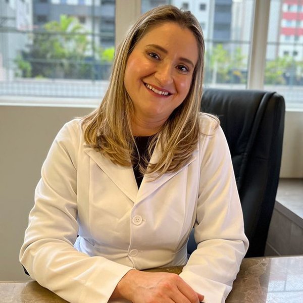 Dra. Simone Zandona Rodrigues, ginecologista, está em seu consultório, com uma ampla janela ao fundo que traz luz natural ao ambiente. Ela está sentada, com as mãos sobre a mesa, sorrindo gentilmente para a câmera com a cabeça levemente inclinada, transmitindo uma atitude acolhedora e atenciosa. Ela tem cabelos lisos e loiros, que caem abaixo dos ombros, e veste uma camisa preta com um jaleco branco por cima, refletindo uma aparência profissional e elegante.