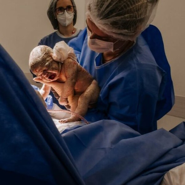 Dra. Simone está no centro cirúrgico logo após realizar um parto, segurando o recém-nascido em seus braços e mostrando-o para a mãe. O bebê, ainda chorando, é acolhido no primeiro contato com o mundo, e o momento captura a emoção e a ternura do nascimento. Duas enfermeiras acompanham a Dra. Simone, completando a equipe que assiste ao parto.
