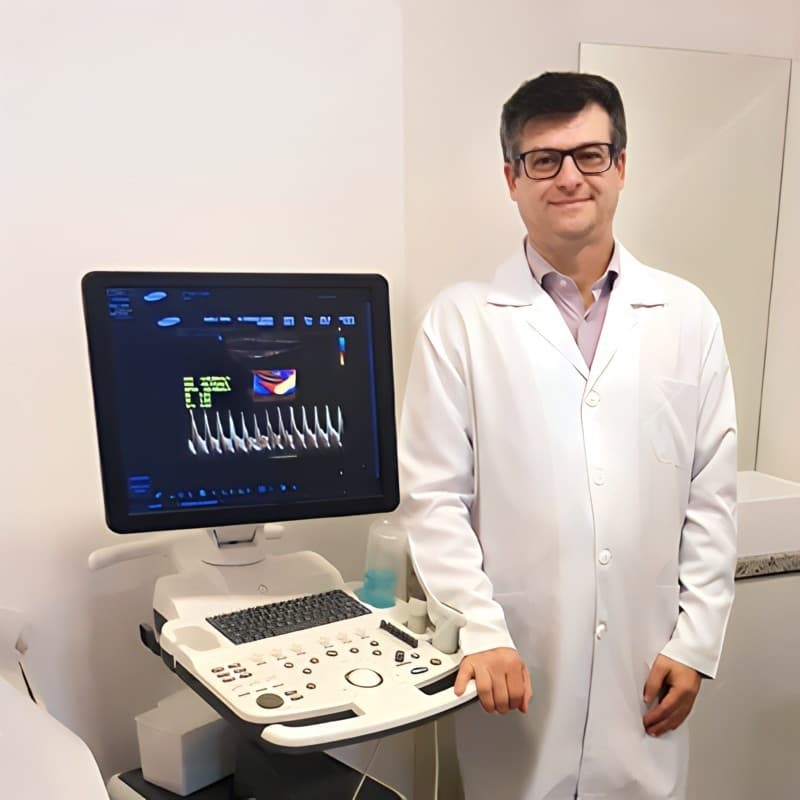 Dr. Rodrigo, cardiologista, olhando para a câmera enquanto está ao lado do seu Ecodoppler que é um dispositivo utilizado para visualizar as veias dos pacientes, ele está vestindo jaleco branco