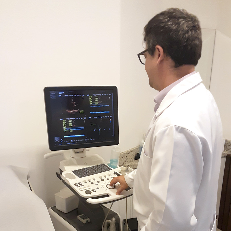 Dr. Rodrigo, cardiologista, de costas para a câmera enquanto está digitando algo em seu Ecodoppler, ele está vestindo jaleco branco