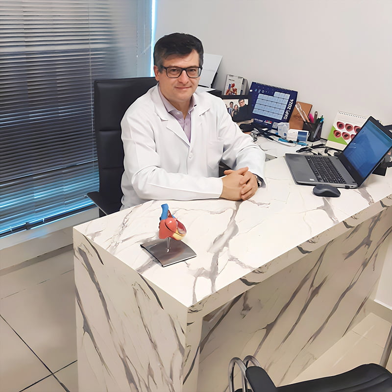 Dr. Rodrigo, cardiologista, sorrindo para a câmera enquanto está com as mãos unidas e sentado em sua mesa de trabalho no consultório, vestindo jaleco branco