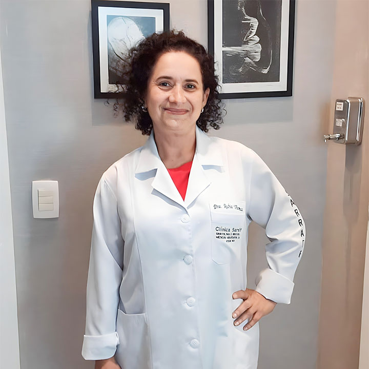 Dra. Rúbia Thomaz de Aquino Antunes em seu consultório, posando com confiança e simpatia. Na imagem, ela está de pé, com uma das mãos na cintura e um sorriso acolhedor dirigido à câmera. Ela veste um jaleco branco com seu nome bordado no bolso, complementado por uma elegante camisa vermelha por baixo. Ao fundo, uma parede cinza emoldura dois quadros exibindo imagens de ultrassonografias gestacionais, conferindo um toque profissional e temático ao ambiente.