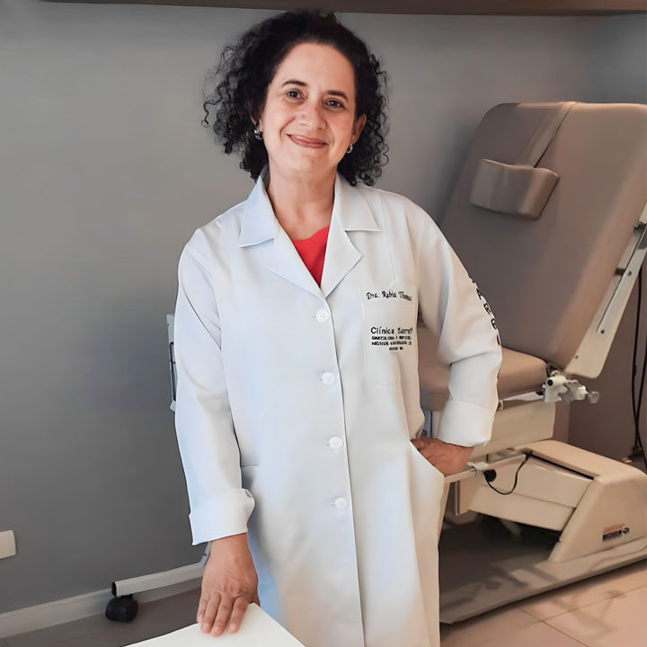 Dra. Rúbia Thomaz de Aquino Antunes em seu consultório, transmitindo simpatia e profissionalismo ao olhar e sorrir para a câmera. Na imagem, ela está de pé, com uma das mãos na cintura e a outra apoiada sobre sua mesa. Ela veste um jaleco branco com seu nome bordado no bolso, reforçando sua identidade profissional. Ao fundo, é possível ver uma cadeira ginecológica, complementando o ambiente médico.