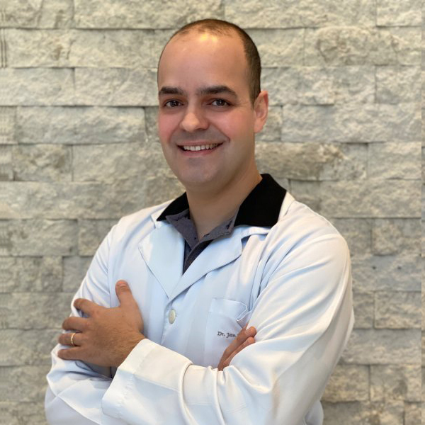 Dr. Jan Pawel aparece olhando diretamente para a câmera com um sorriso simpático e acolhedor. De braços cruzados, veste um jaleco branco. O fundo da imagem apresenta uma parede decorativa com tijolos de pedra em tons de bege.