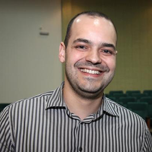 Dr. Jan Pawel aparece olhando diretamente para a câmera com um sorriso simpático. Ele veste uma camisa listrada branca e preta. O fundo da imagem apresenta um cenário que lembra uma sala de convensões com muitas cadeiras.