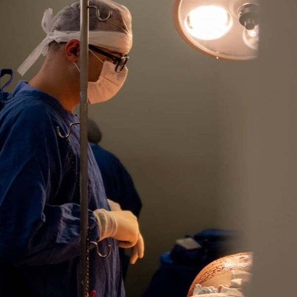 Dr. Lucas Marin Dall Stella está no centro cirúrgico, concentrado enquanto examina o abdômen de uma paciente recém-operada, momentos após o término da cirurgia. Sua expressão reflete seriedade e dedicação ao cuidado com a paciente.