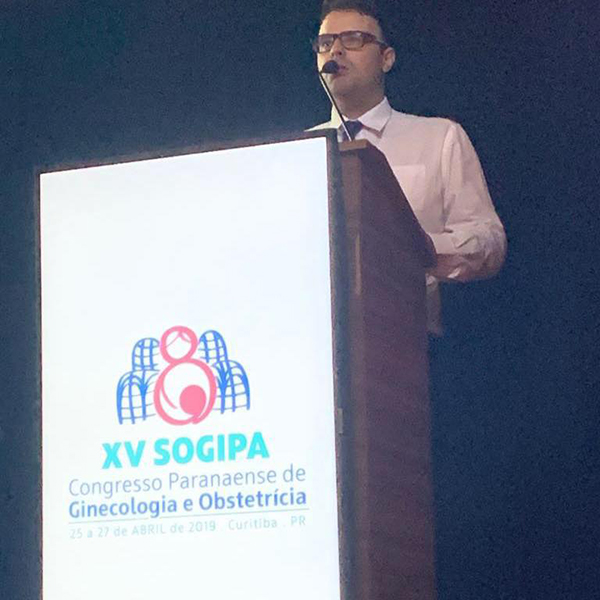 Dr. Lucas Marin Dall Stella discursa no Congresso Paranaense de Ginecologia e Obstetrícia. Com um microfone em mãos, ele está posicionado atrás de um púlpito, dirigindo-se a uma plateia de ginecologistas atentos. Sua postura reflete confiança e domínio sobre o tema apresentado.