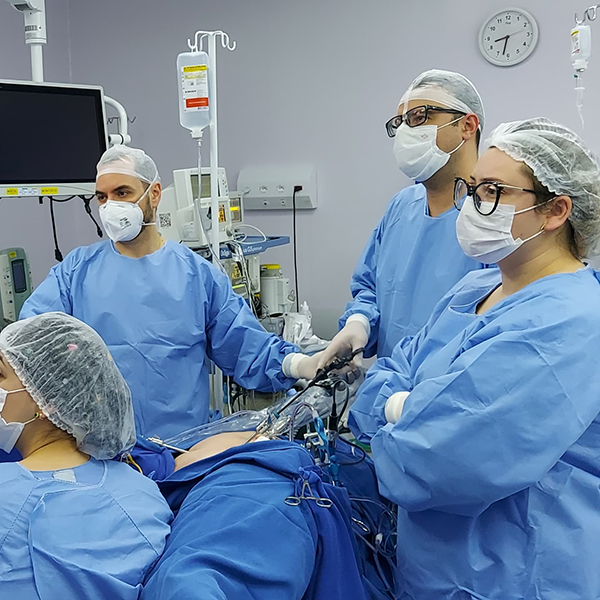 Dr. Lucas Marin Dall Stella conduz uma cirurgia por vídeo no abdômen de uma paciente, no centro cirúrgico. Ele está acompanhado por três auxiliares, todos vestidos com jalecos azuis. A equipe demonstra seriedade e concentração enquanto realiza o procedimento.