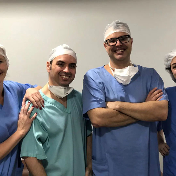 Dr. Lucas Marin Dall Stella está no centro, cercado por três auxiliares de sua equipe cirúrgica. Ele é o mais alto, com os braços cruzados, destacando-se no meio do grupo. Todos estão vestidos com jalecos azuis e olham para a câmera com sorrisos que transmitem confiança e simpatia.