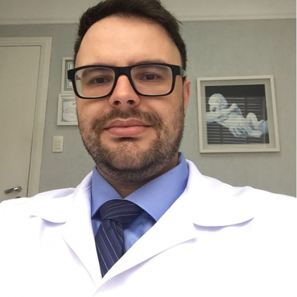 Dr. Lucas Marin Dall Stella está em seu consultório, olhando diretamente para a câmera em um close. Vestindo jaleco branco, camisa azul e gravata, ele exibe uma expressão séria e profissional.