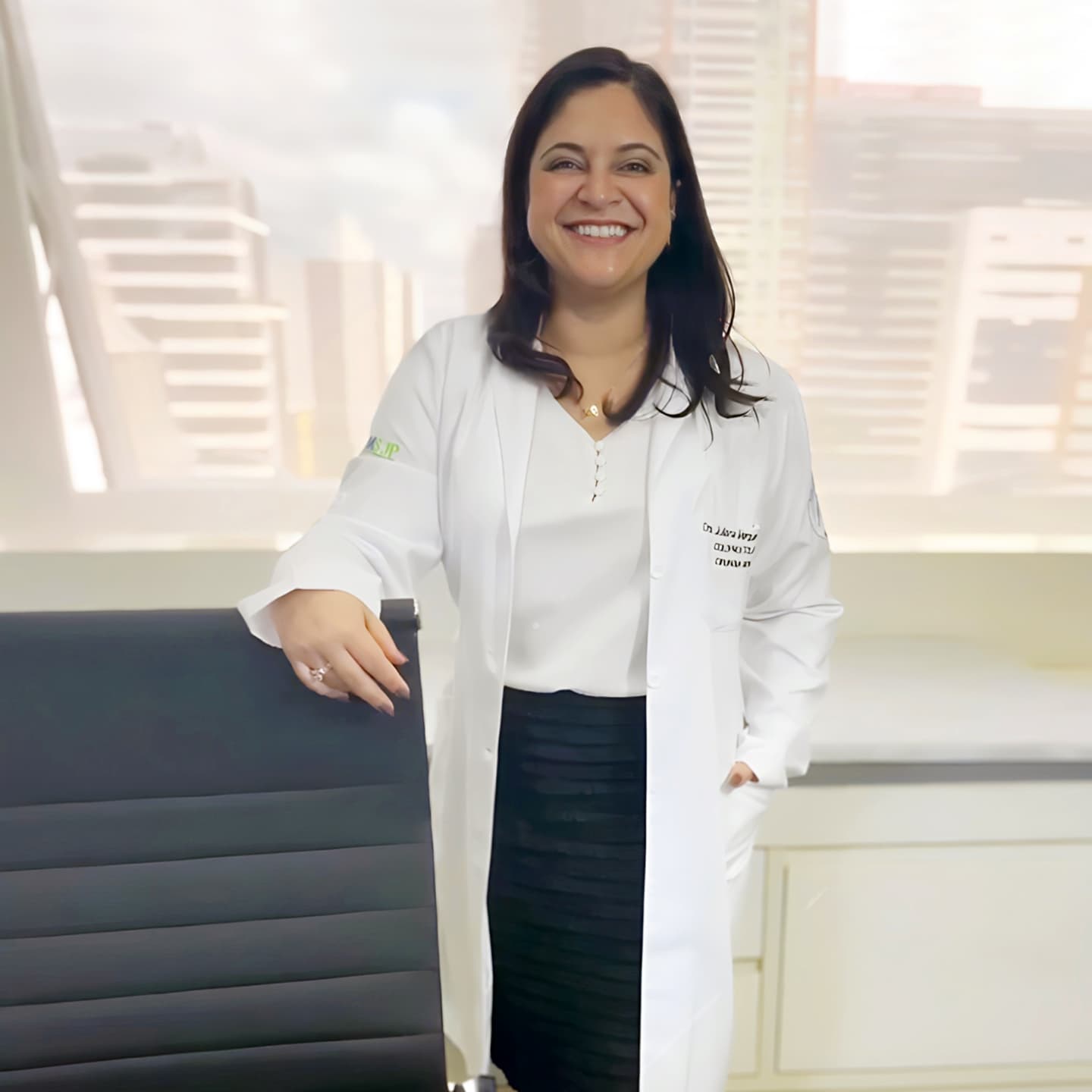 Dra. Juliana, coloproctologista, sorrindo para a câmera enquanto está de de pé ao lado da sua cadeira de trabalho no consultório, vestindo saia preta, camisa branca e jaleco branco.