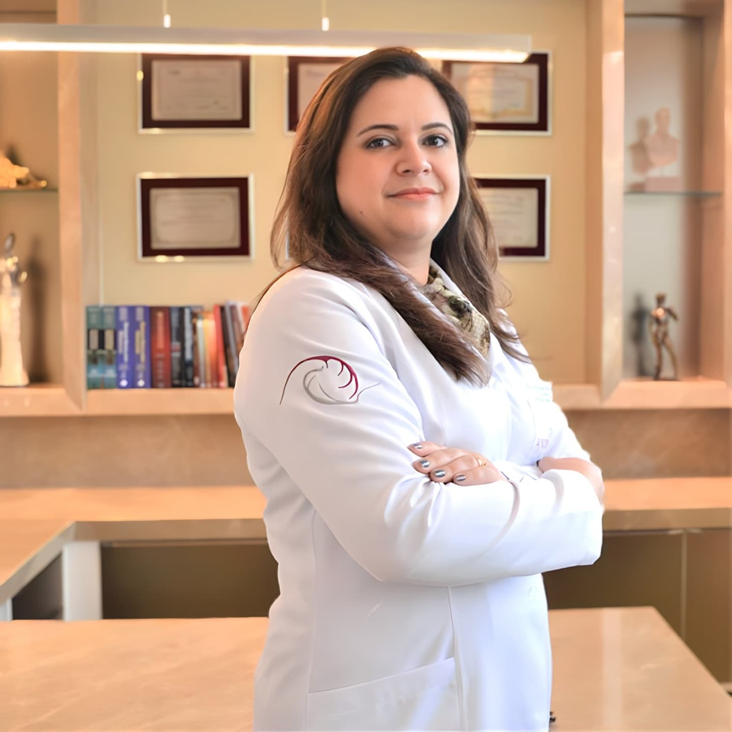 Dra. Juliana, coloproctologista, sorrindo discretamente para a câmera enquanto está de pé e com os braços cruzados em consultório, ela está vestindo jaleco branco.