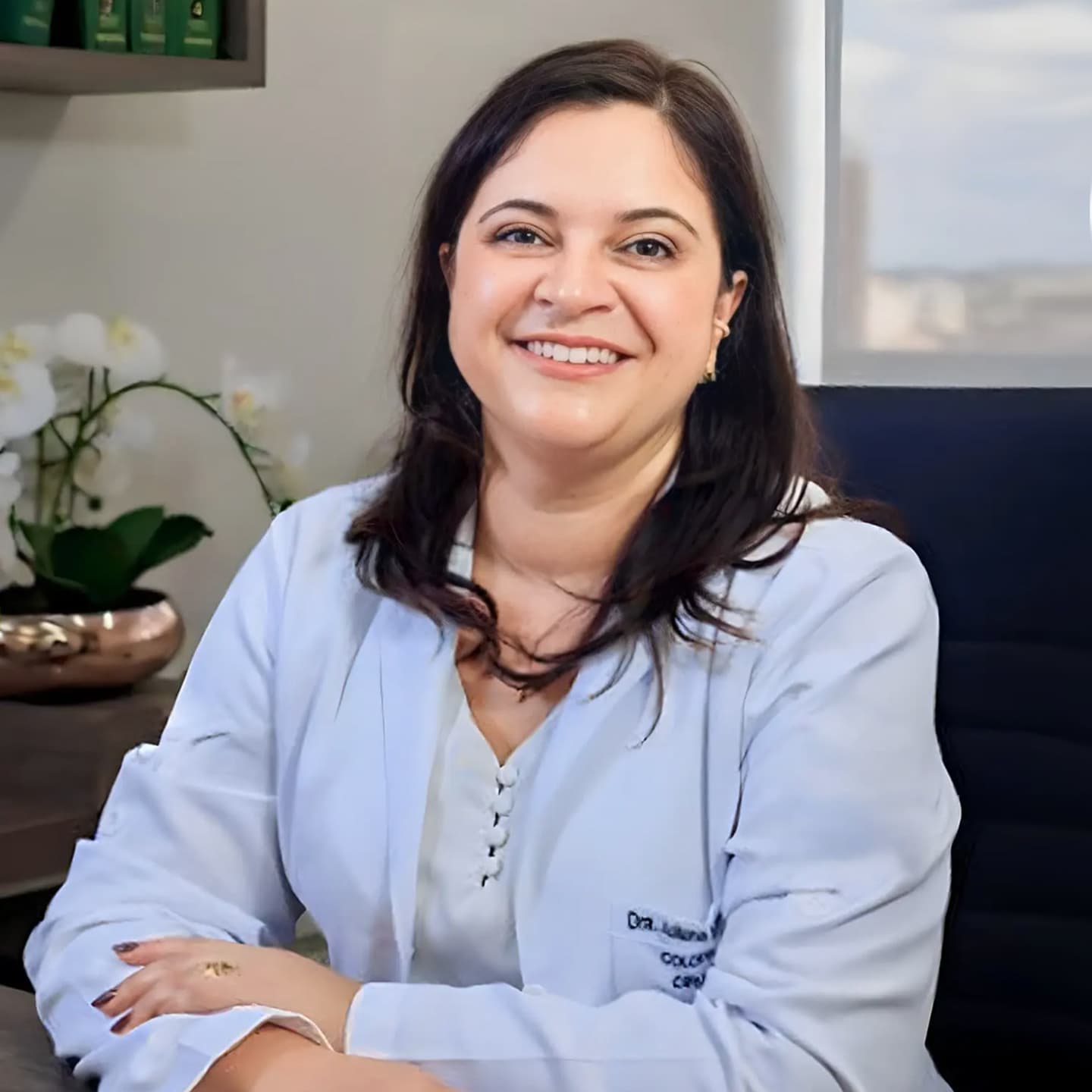 Dra. Juliana, coloproctologista, sorrindo para a câmera enquanto está sentada em sua mesa de trabalho no consultório, vestindo camisa branca e jaleco branco.