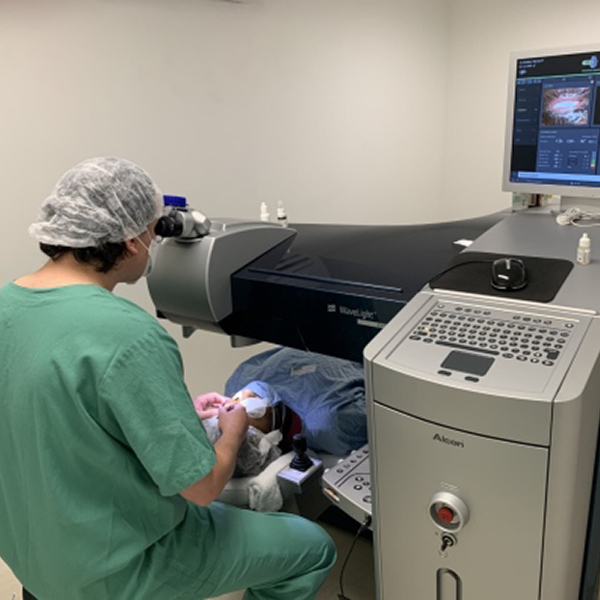 Dr. Fernando vestindo roupa cirúrgica sentado em uma cadeira operando uma máquina de cirurgia laser pela qual está realizando uma cirurgia oftalmológica em um paciente deitado por debaixo dessa máquina.