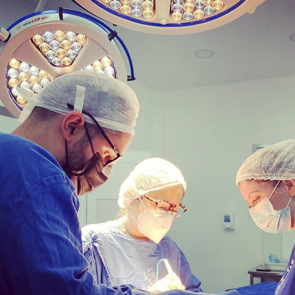 Dr. João Abelha com roupa cirúrgica fazendo cirurgia em centro cirúrgico, acompanhado de duas assitentes.