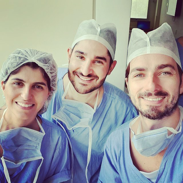 Dr. João Abelha com roupa cirúrgica em centro cirúrgico acompanhado de dois colegas, todos olhando e sorrindo para a câmera.