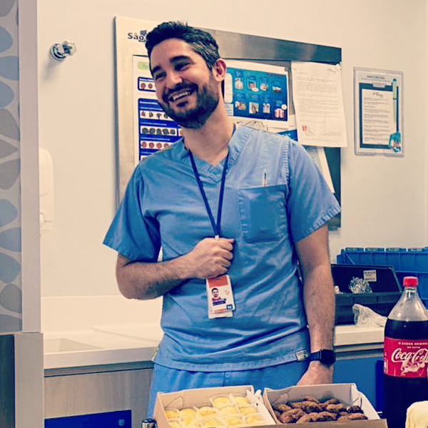 Dr. João Abelha com roupa cirúrgica sorrindo e olhando para o lado em uma sala com refrigerante e salgadinhos sobre uma mesa.