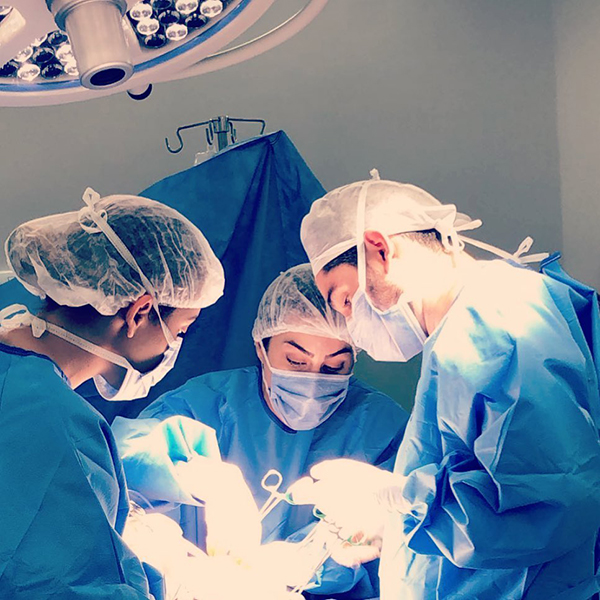 Dr. João Abelha com roupa cirúrgica fazendo cirurgia em centro cirúrgico, acompanhado de duas assitentes.