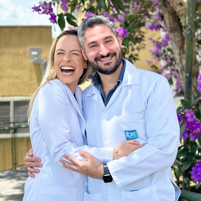 Dr. João Abelha abraçado com uma colega e ambos sorrindo muito, eles estão com roupa branca de médico e em um lugar ao ar livre.