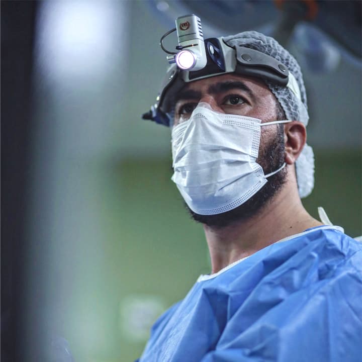 Dr. Mohamad, na sala de cirurgia olhando para o monitor durante a cirurgia. Ele está com barba e máscara cirúrgica.