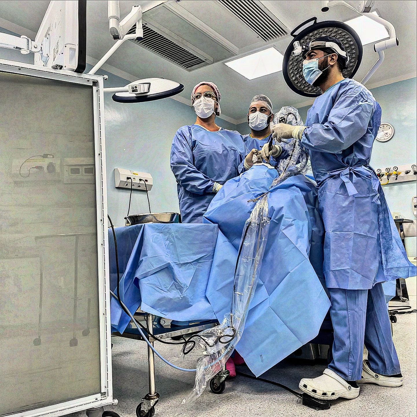 Dr. Mohamad, na sala de cirurgia olhando para o monitor durante a cirurgia. Ele está com barba e máscara cirúrgica.