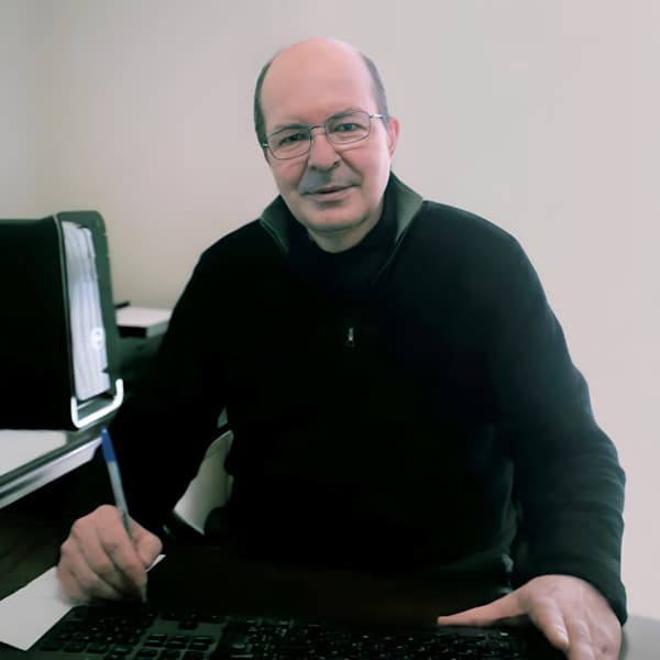 Foto do Dr. Rui Max Beckhauser, Psiquiatra em Curitiba. Ele está sentado em sua mesa de trabalho, usando óculos e uma blusa escura, com uma expressão acolhedora e profissional. Ele segura uma caneta na mão, enquanto parece revisar ou assinar documentos. Ao fundo, é possível ver um computador e detalhes do ambiente que sugerem um consultório ou espaço de trabalho organizado.