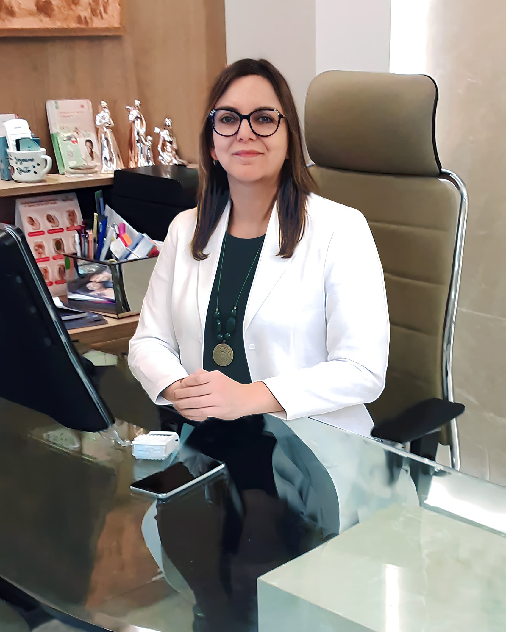 Dra. Joseane Aparecida de Brito, ginecologista em Curitiba, meia idade, cabelo channel liso e loiro médio, muito simpática olhando e sorrindo para a câmera, sentada em sua poltrona aparentemente em seu consultório. ela está de óculos e vestindo uma camiseta preta com um jaleco branco por cima, por trás uma parede em mármore bege.