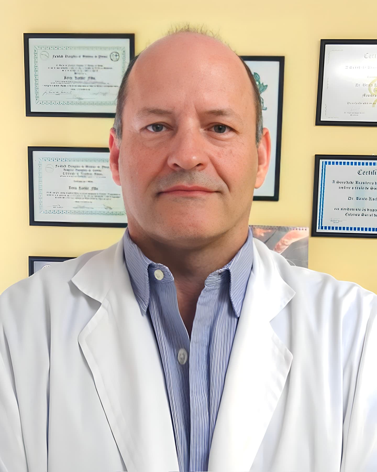 Dr. Bento Koehler Filho, ortopedista em Curitiba, meia idade, pouco cabelo curto e castanho, levemente sorrindo e olhando para a câmera, ele está de pé com uma parede por trás com quadros de diplomas, ele está usando camisa listrada azul com jaleco branco por cima.