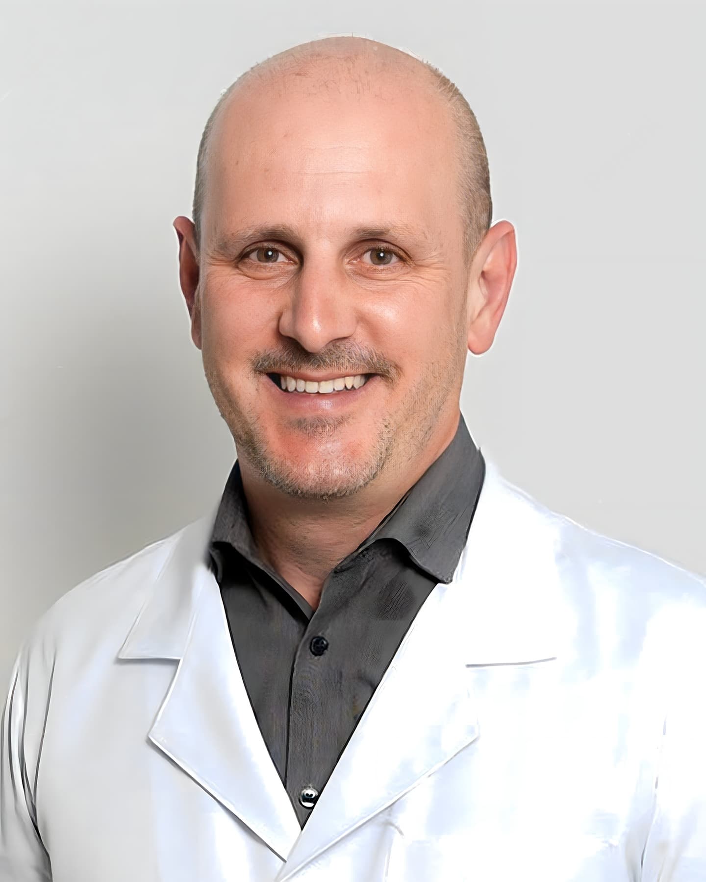 Dr. Marcelo Butenas Piluski, ortopedista de meia-idade, está em pé, com um sorriso enquanto olha diretamente para a câmera. Ele exibe uma aparência profissional, com cabelos levemente ralos, vestindo um jaleco branco sobre uma camisa cinza.