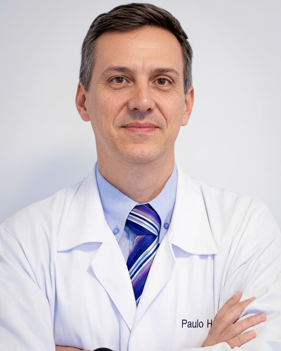 Dr. Paulo Henrique Stahlke, cirurgião vascular em Curitiba, meia idade, cabelos curtos castanhos, de braços cruzados, olhando e sorrindo discretamente para a câmera enquanto está de pé, ele está vestindo um jaleco branco, por trás uma parede branca.