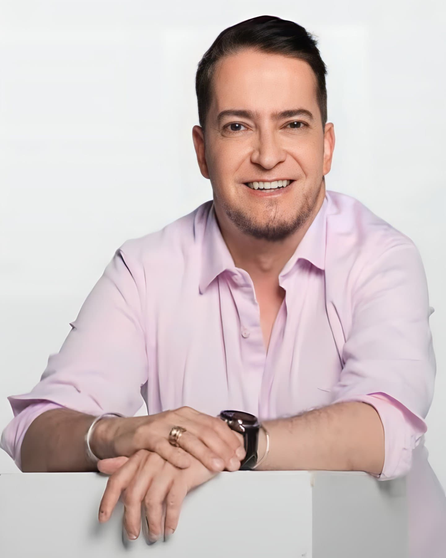Dr. Francisco Germiniani, neurologista em Curitiba, meia idade, cabelos castanhos médio e curtos, barba castanha sem bigode, sorrindo e olhando para a câmera, com as mãos juntas e os braços apoiados sobre um balcão, ele está usando uma camisa rosa, óculos e pulseiras, por trás uma parede cinza claro.
