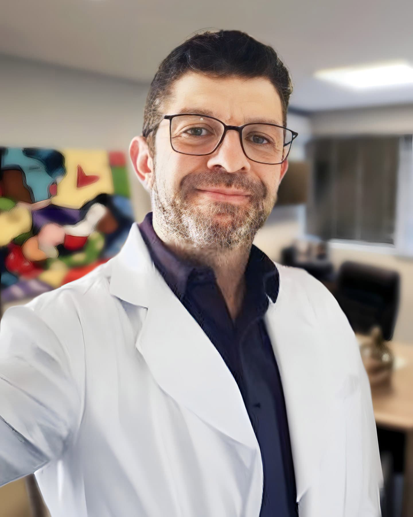 Dr. Marcelo Guimarães Rodrigues, ginecologista, aparece em seu consultório, olhando para a câmera com um sorriso acolhedor. Ele tem cabelos curtos castanho-escuros, uma barba rala e usa óculos, transmitindo uma imagem simpática e profissional. Ele está vestido com uma camisa azul-marinho e um jaleco branco por cima. Ao fundo, em desfoque, vê-se seu consultório moderno e acolhedor.