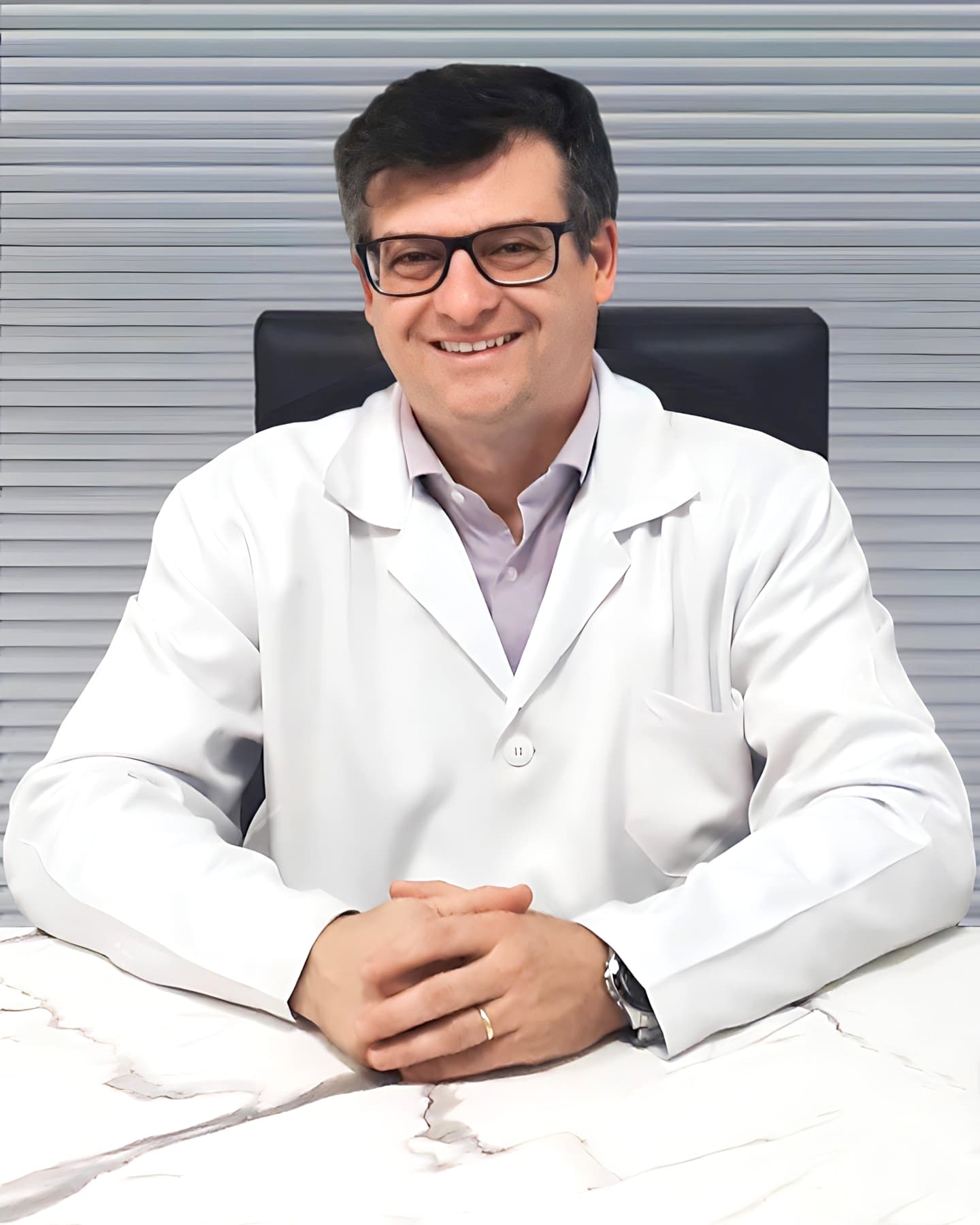 Dr. Rodrigo Lazzari, cardiologista em Curitiba, meia idade, cabelos curtos e castanhos, sorrindo para a câmera enquanto está sentado, com os braços sobre sua mesa de trabalho no consultório, com as mãos juntas, ele está usando óculos, relógio e está vestindo uma camisa lilás claro com um jaleco branco por cima, por trás uma veneziana cinza fechada.