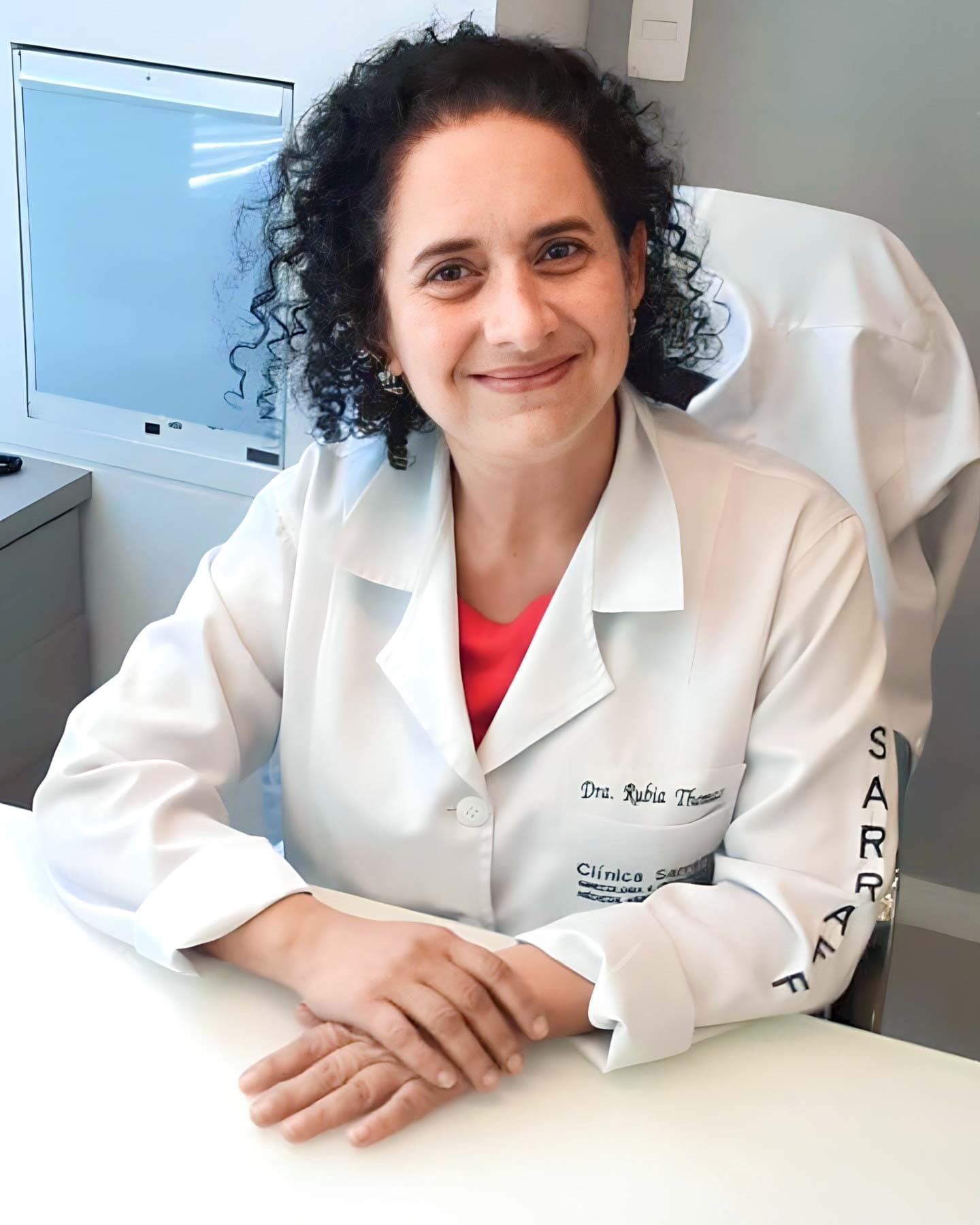 Dra. Rúbia Thomaz de Aquino Antunes é ginecologista com consultórios em Pinhais e Curitiba. De meia-idade, possui cabelos castanhos encaracolados que caem até os ombros. Na imagem, ela está sentada em sua mesa de consultório, com as mãos gentilmente sobrepostas sobre a superfície da mesa. Seu olhar é direcionado à câmera, acompanhado por um sorriso acolhedor. Ela veste um jaleco branco, que traz seu nome bordado no bolso, e por baixo do jaleco usa uma elegante camisa vermelha.