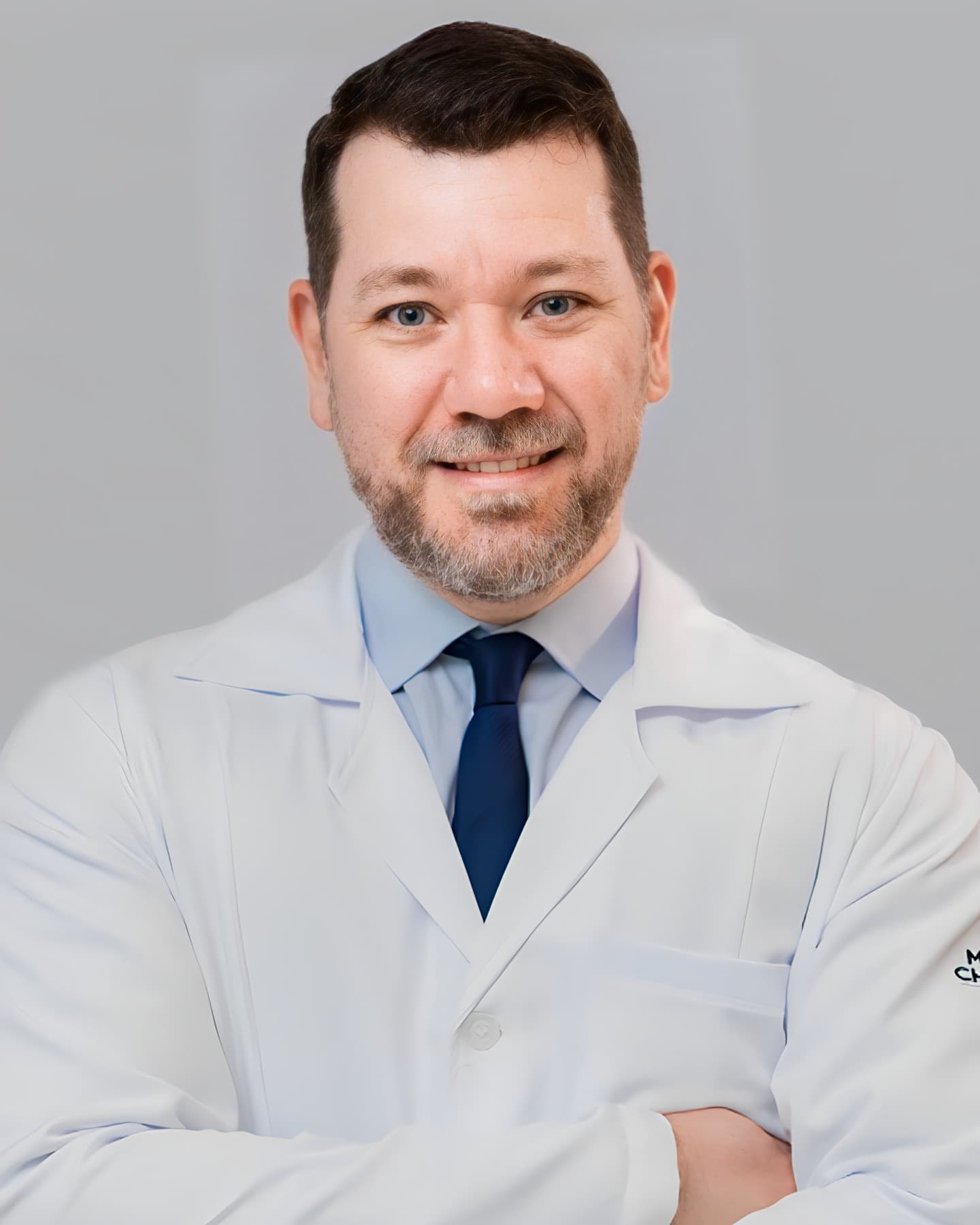 Dr. José Augusto Ribas Fortes, cardiologista em Curitiba, meia idade, cabelos curtos castanhos, de braços cruzados e sorrindo para a câmera, trajando gravata zaul, camisa azul claro e um jaleco branco por cima, por trás uma parede cinza.