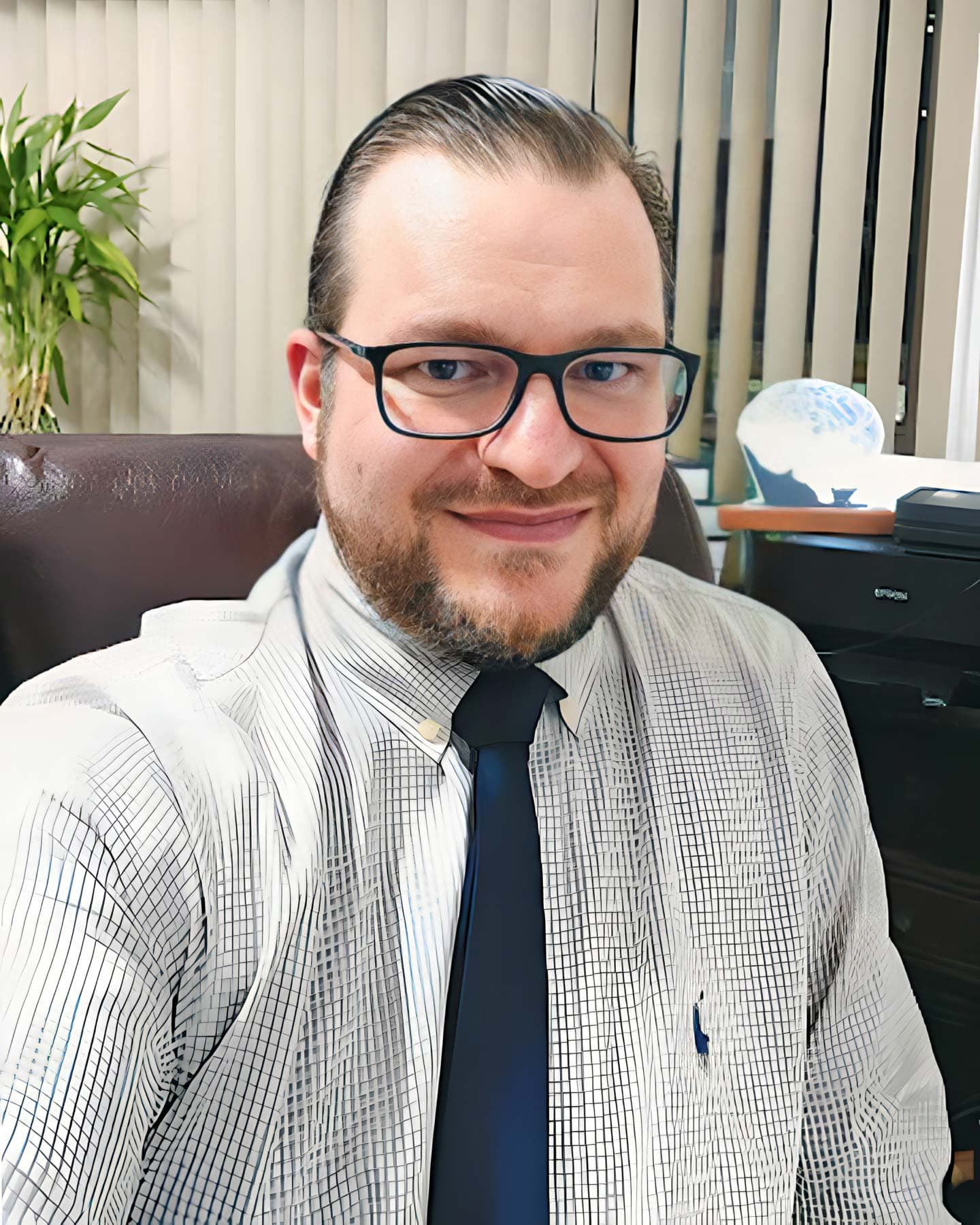 Dr. Ricardo Manzochi Assmé, psiquiatra em Curitiba, meia idade, cabelo lisos curtos castanho médio com gel penteado pra trás, barba bem feita castanho médio, sorrindo discretamente e olhando para a câmera, ele está sentado aparentemente em seu consultório, por trás uma janela com cortinas verticais bege, uma planta e uma impressora preta.