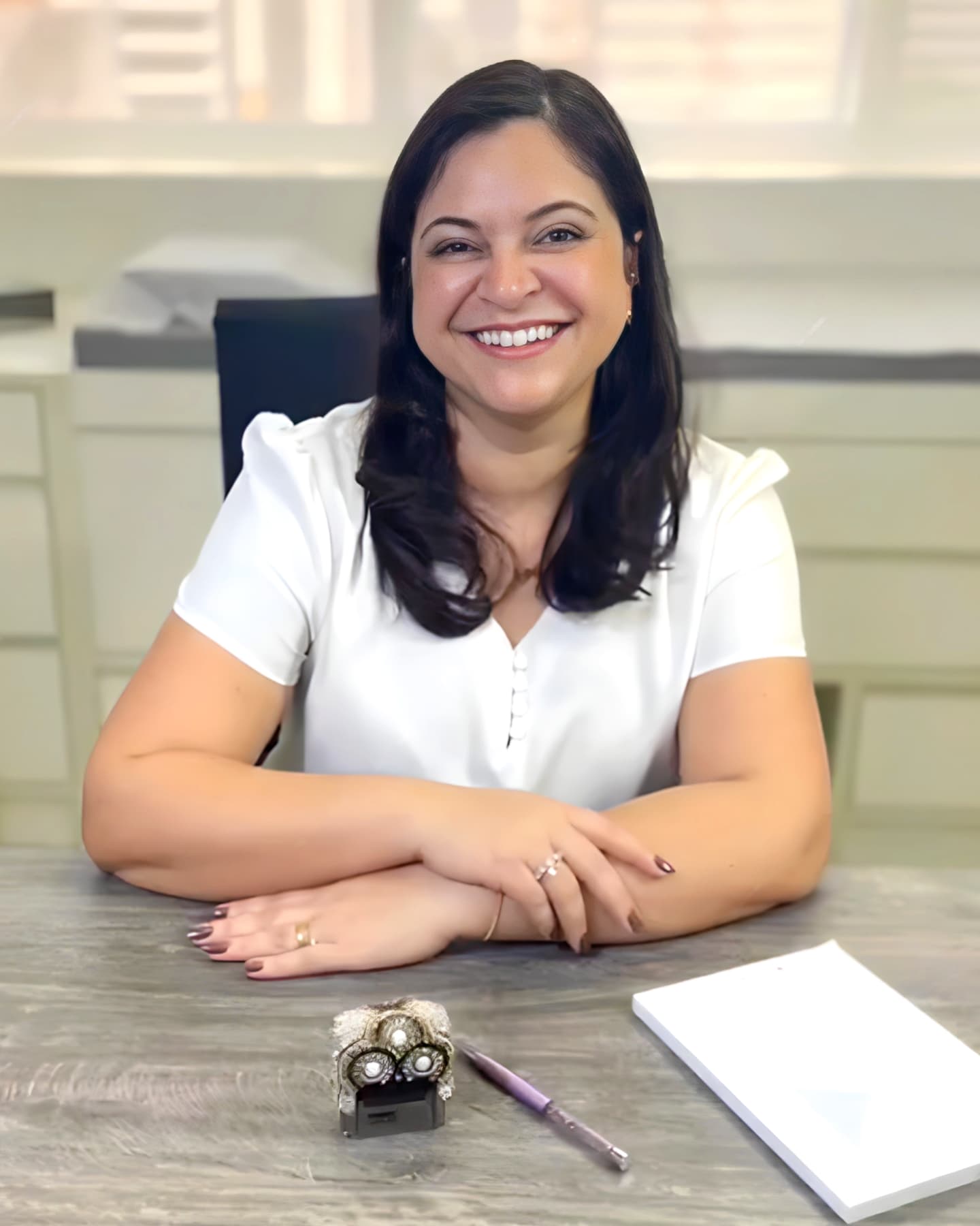 Dra. Juliana Marques Ferreira, coloproctologista em Curitiba, jovem de meia idade, cabelos longos, lisos e castanhos claro, sorrindo para a câmera enquanto está sentada em sua mesa de trabalho no consultório, apoiando os braços sobre a mesa, ela está vestindo uma camisa branca, por trás está a maca utilizada para exames e procedimentos.