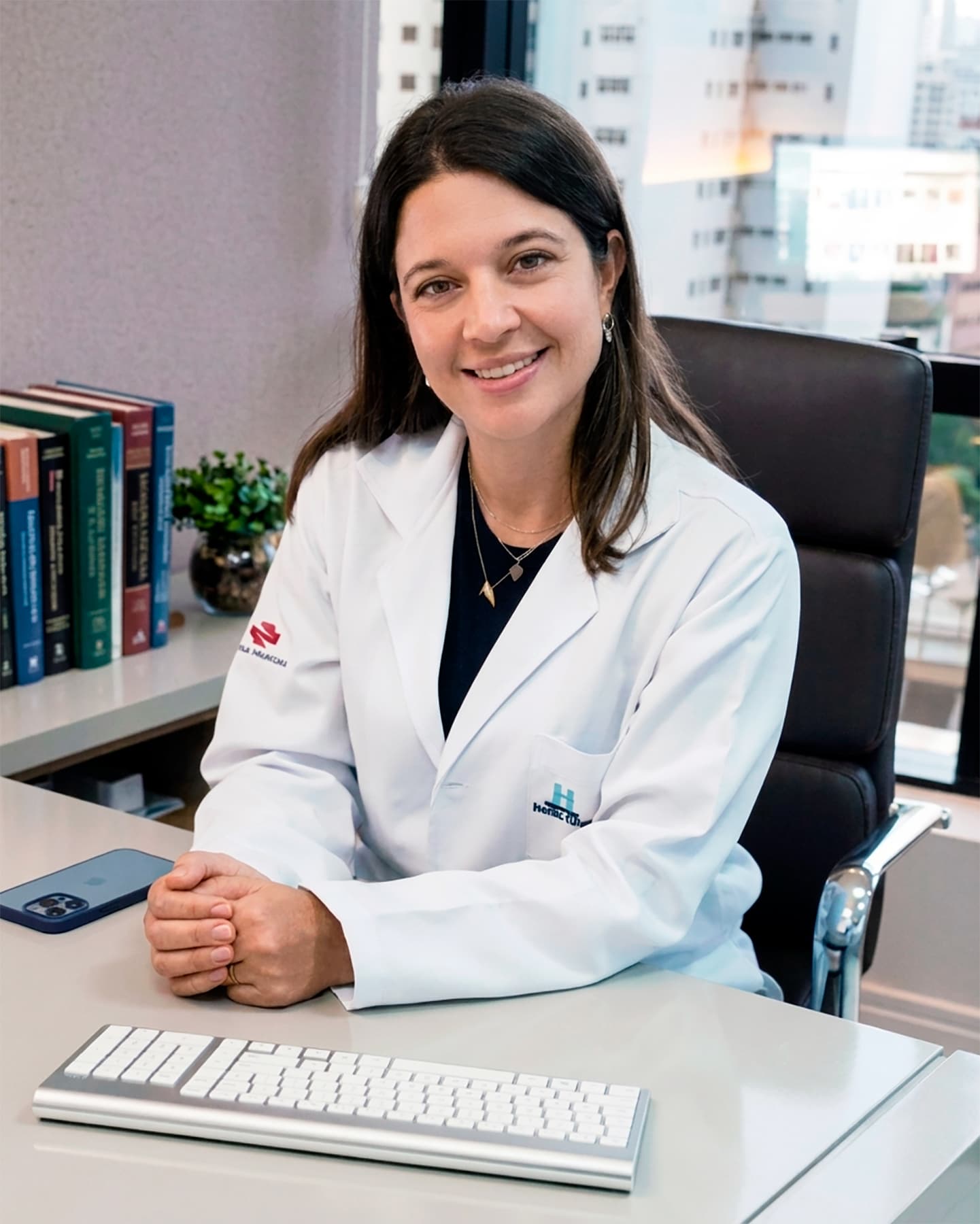 Dra. Mariana Drechmer Romanowski, médica de meia-idade, olha para a câmera com um sorriso acolhedor. Seus cabelos castanhos médios, lisos e na altura dos ombros, emolduram seu rosto de forma elegante. Ela está sentada em seu consultório, com as mãos juntas e sobre a mesa. Ela veste um jaleco branco com uma camisa preta por debaixo, que combina com o ambiente profissional e moderno de seu consultório. Ao fundo, uma ampla janela que ocupa toda a parede, permitindo a entrada de luz natural. O consultório transmite uma sensação de conforto e sofisticação, refletindo sua dedicação em proporcionar um espaço acolhedor para suas pacientes.