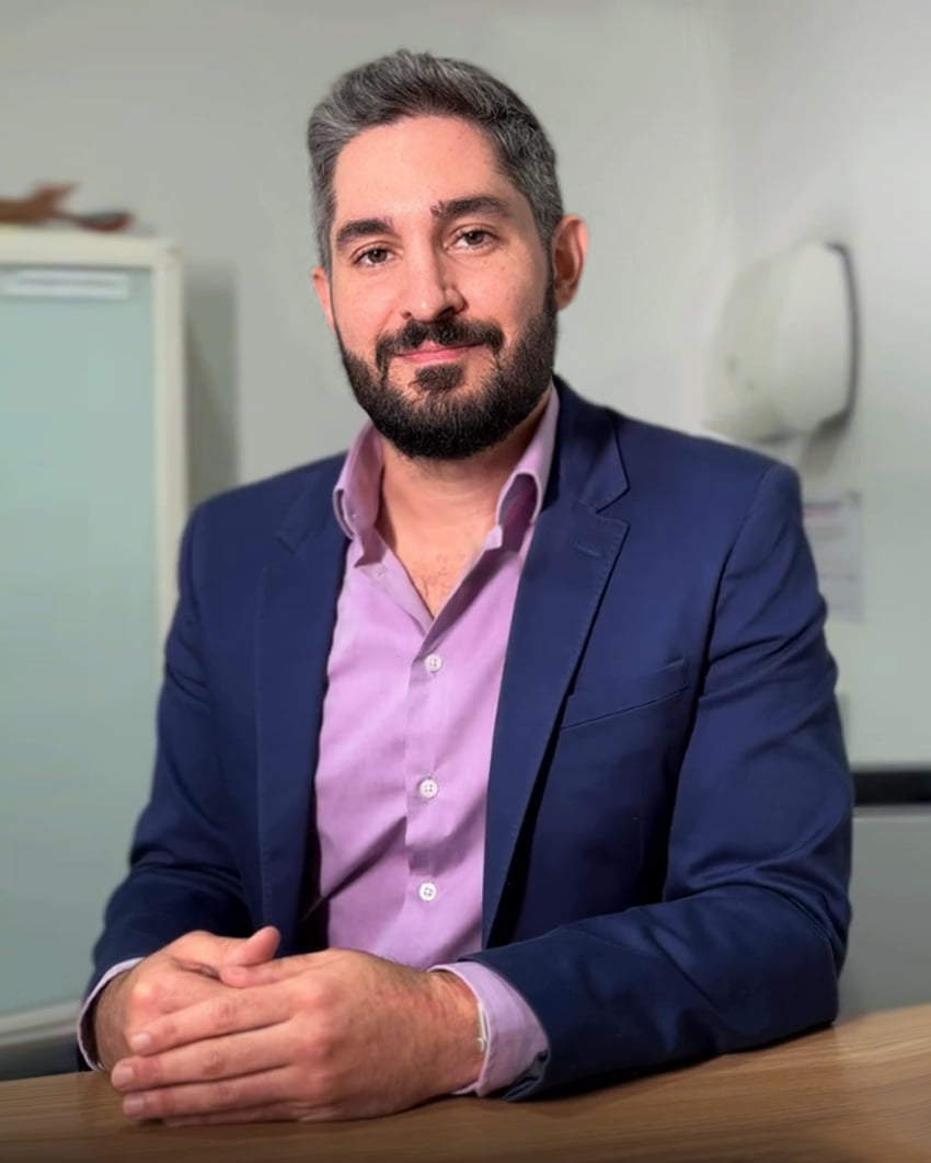 Dr. João Abelha, mastologista e ginecologista, jovem, cabelos curtos castanhos e barba castanha, muito simpático sorrindo para a câmera, está de pé abraçado com uma jovem, ambos estão aprentemente em um centro cirúrgico, ambos vestem toucas brancas e roupas azuis cirúrgicas.