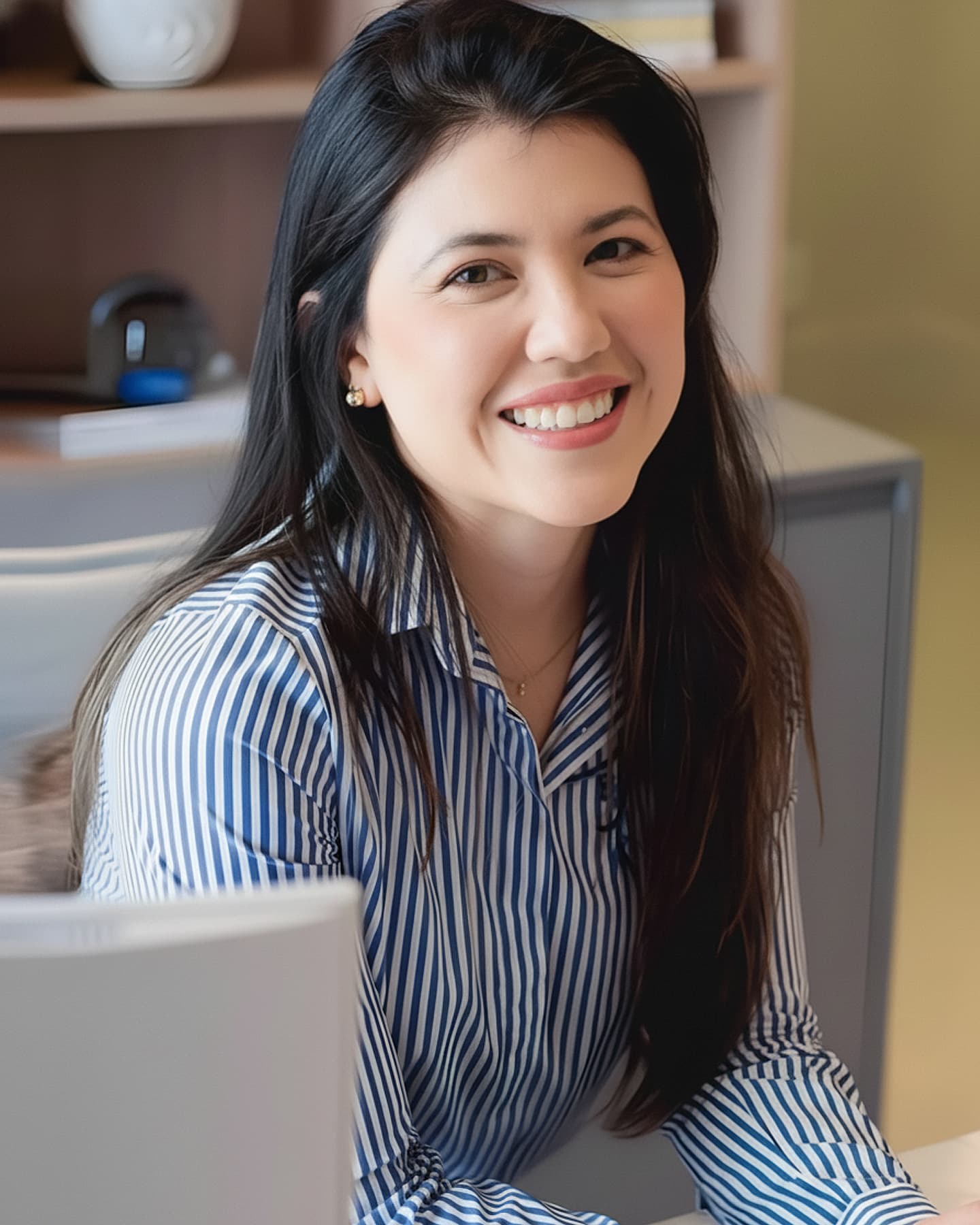 Dra. Ariane Carla Horiuchi, Ginecologista, jovem, cabelos longos castanhos escuros, sentada com seus braços apoiados sobre sua mesa em seu consultório, olhando e sorrindo para a camera. Ela está usando uma camisa listrada branca e azul. Ao fundo, um armário com decoração profissional.